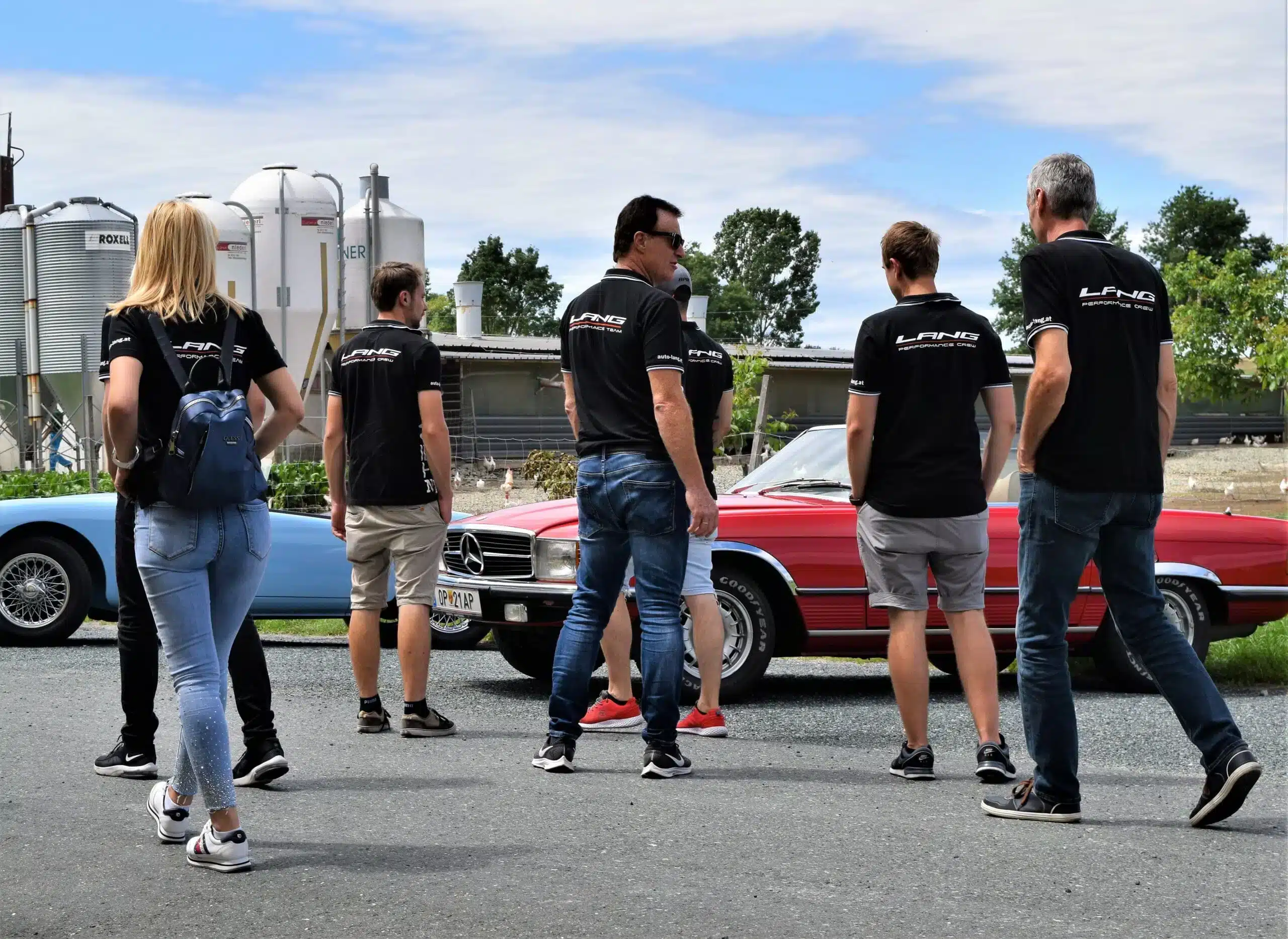 7 Mitglieder des Auto-Lang Teams gehen gemeinsam an Fahrzeugen vorbei. Die Teamkleidung ist ein schwarzes T-Shirt mit der Aufschrift "Lang Performance" in weiß.
