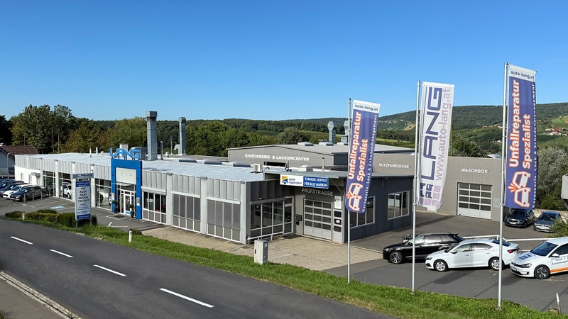 Eine Autowerkstatt mit Schauraum und einer Lackierkabine. Am Eingang steht groß "LANG", mehrere Fahnen sind mit LANG" und "Unfallreparaturspezialist" beschriftet.