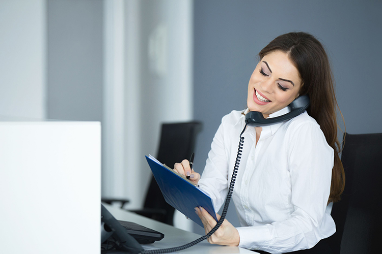 Eine dunkelhaarige Frau sitzt mit einem Notizzettel und einem Telefonhörer in einem Büro