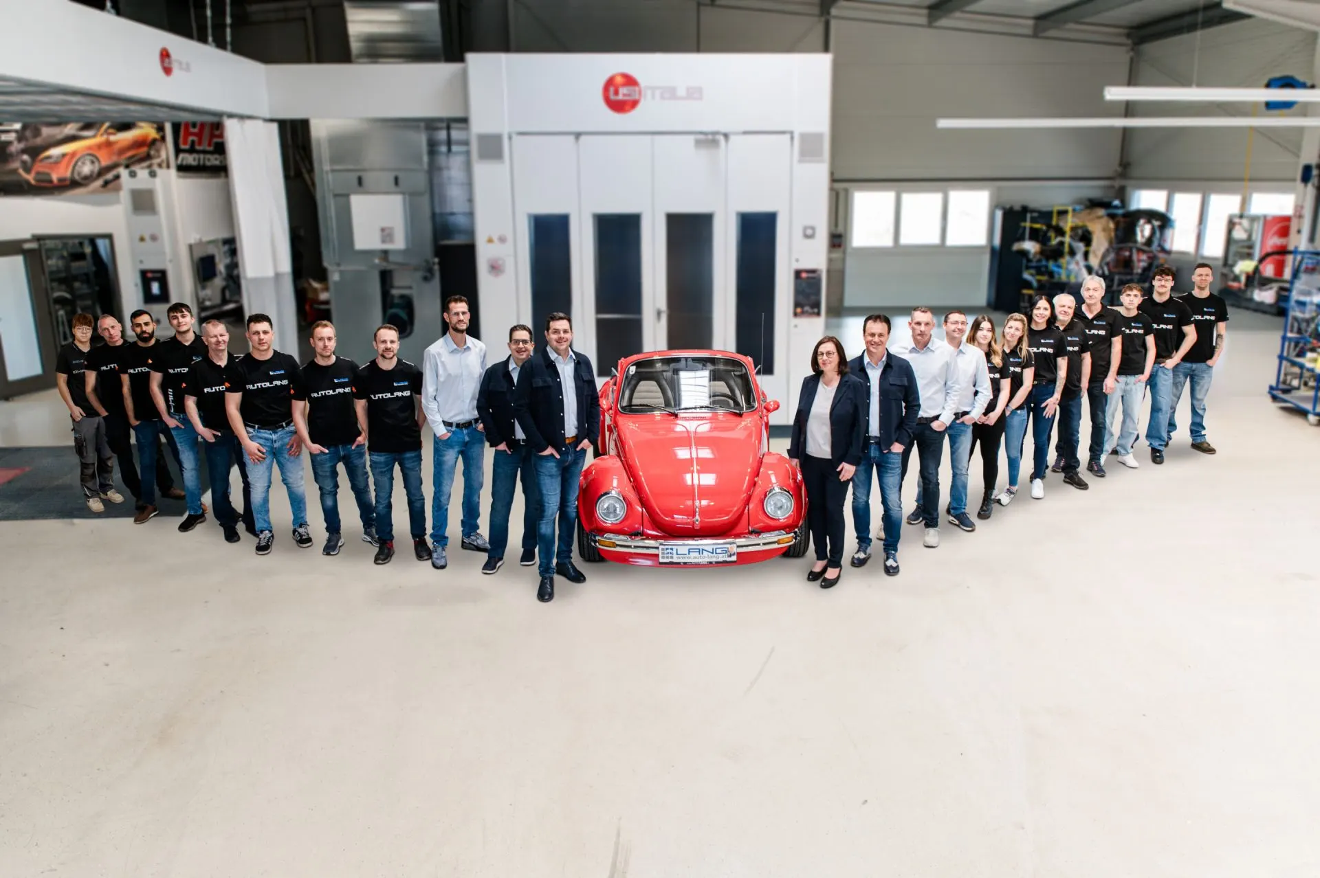 Das Autohaus Lang Team steht in der Lackier- und Karosserieabteilung versammelt um einen roten VW Käfer herum
